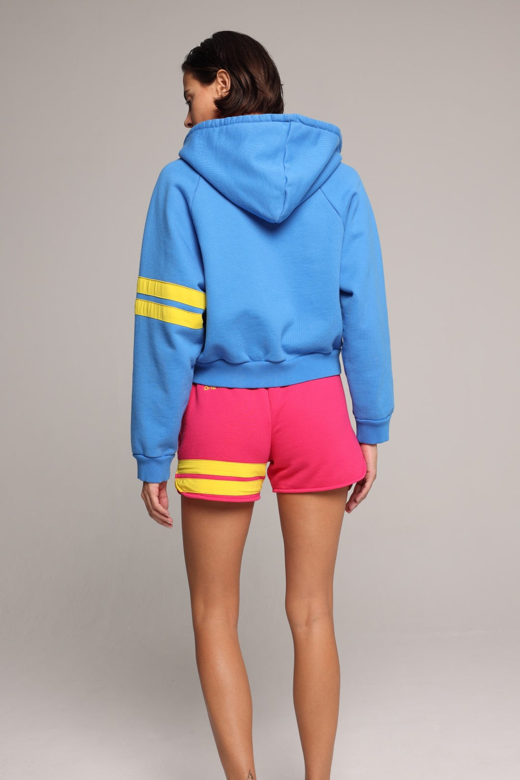 A woman shown from the back wearing a bright blue hoodie with yellow stripes on the sleeves and hot pink shorts with yellow stripes near the hem, against a plain light gray studio background. Premium Sportswear by One More Reason.